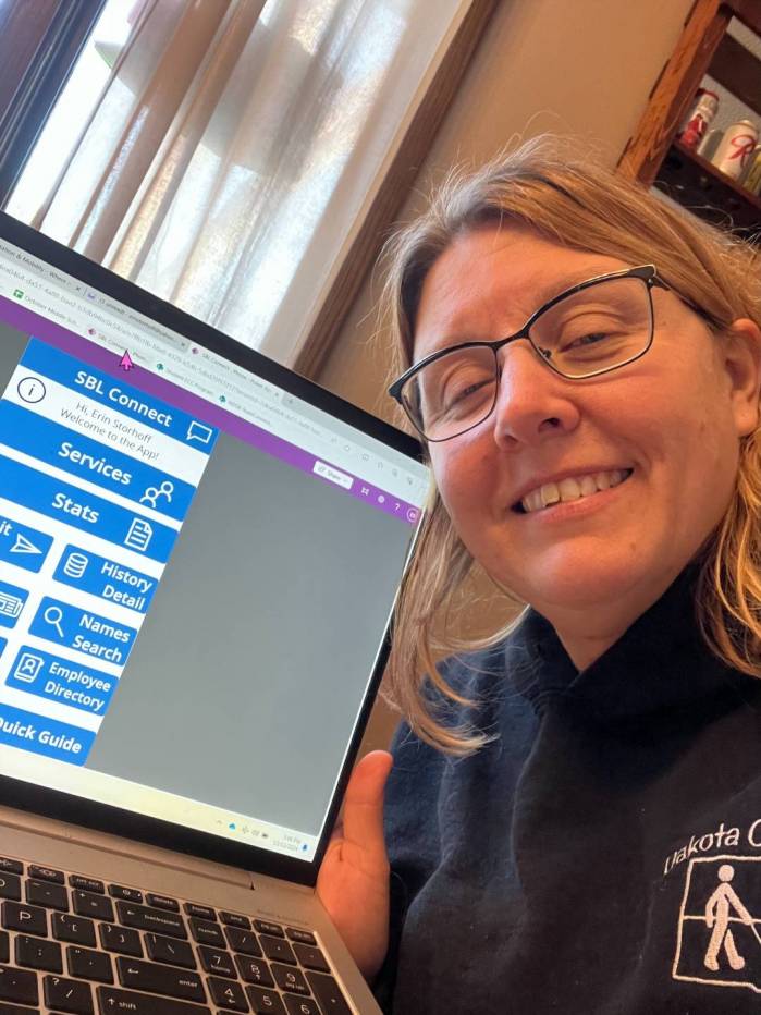 Erin smiles as she holds her laptop computer open to the camera while working on reports in her home.